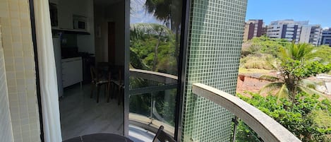 A balcony with a table and chairs, a view of a lush garden, and a glimpse of a kitchen inside.