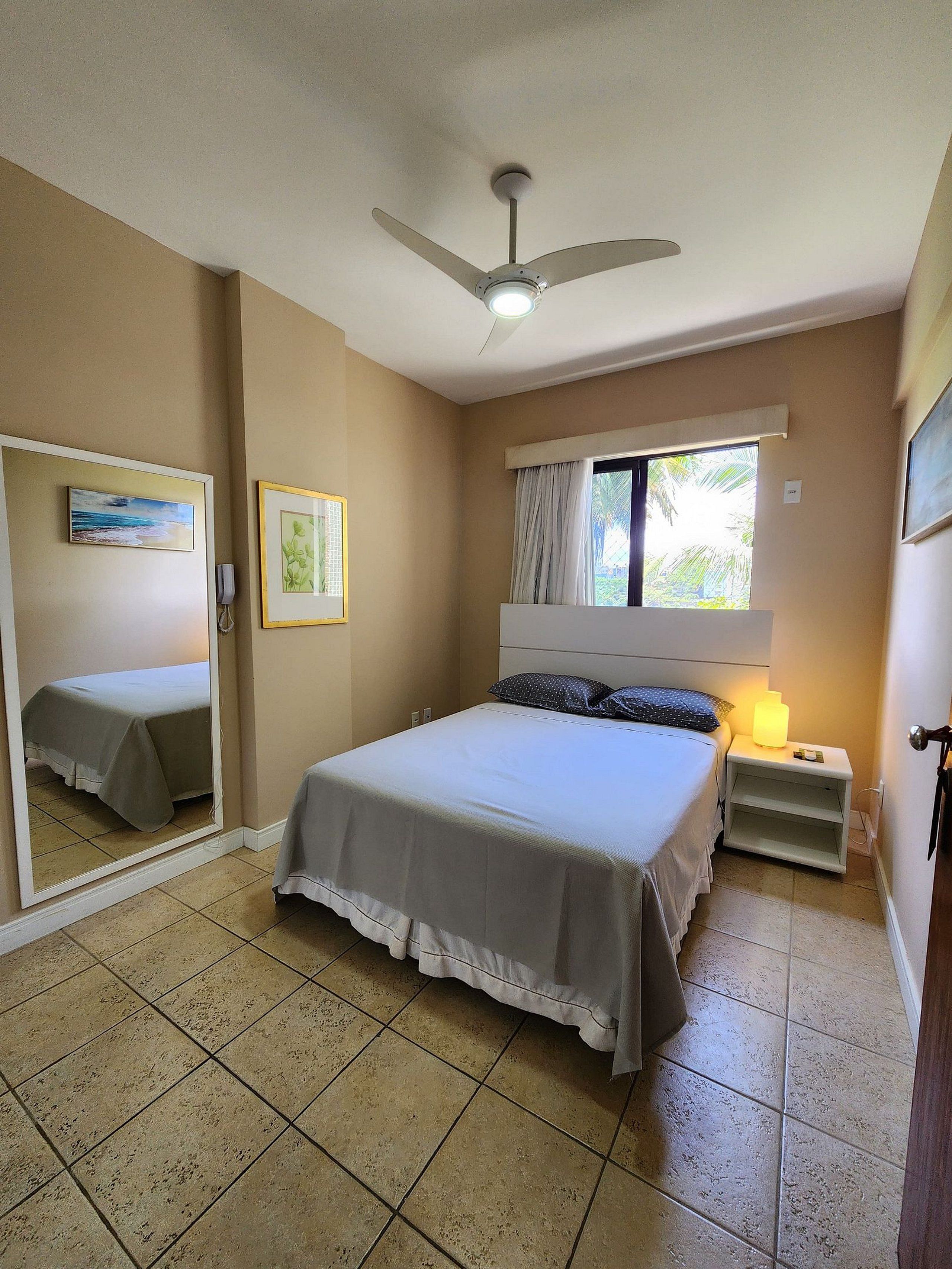 A bedroom with a bed, a ceiling fan, a bedside table with a lamp, a window with curtains, and a mirror on the wall.