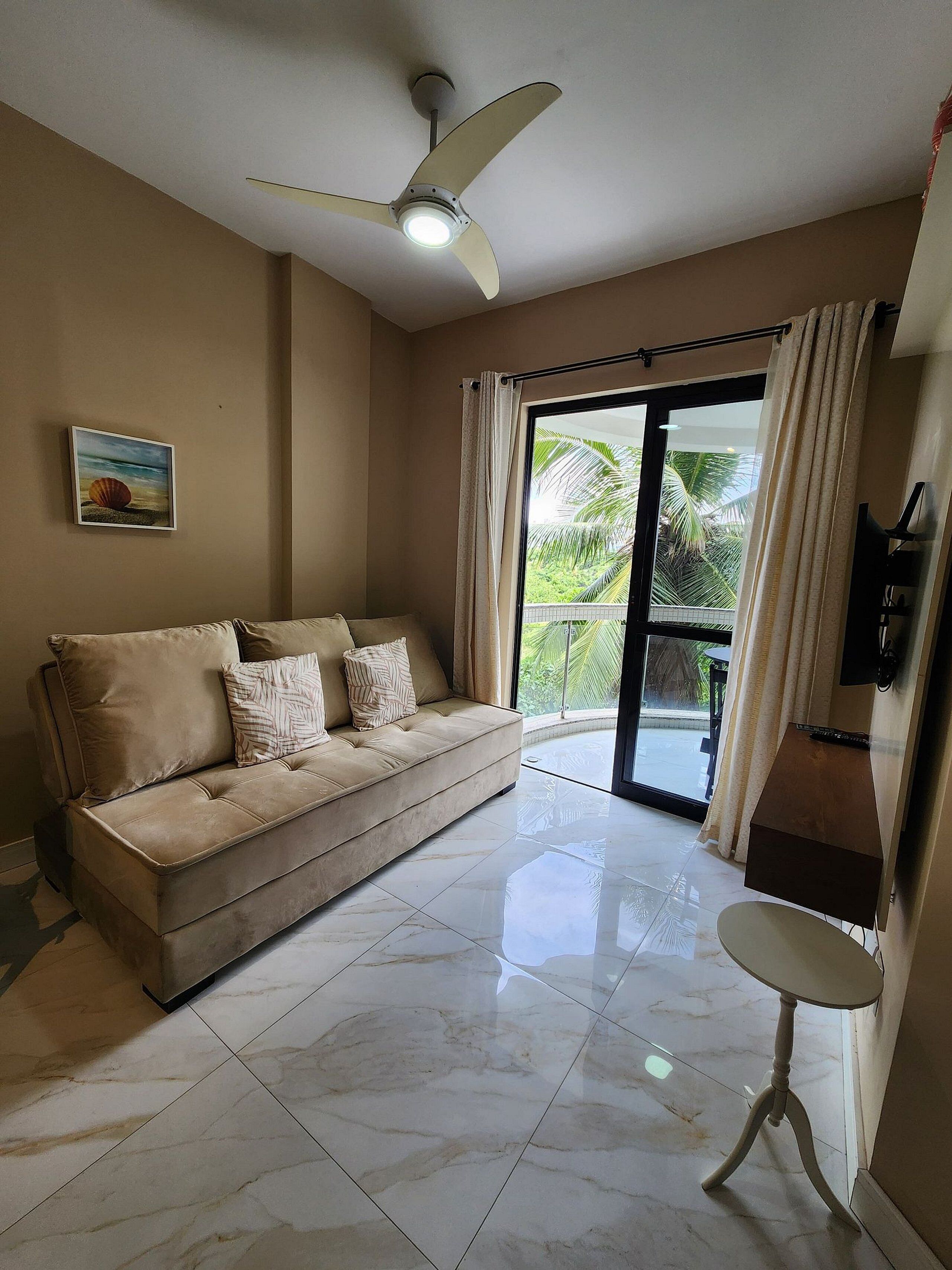 A living room with a beige sofa, a ceiling fan, a painting, and a view of greenery through a glass door.