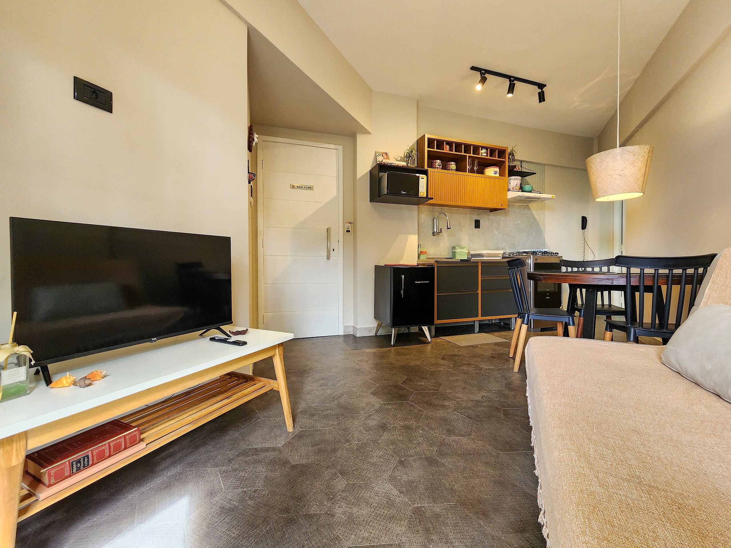 A compact living space with a flat-screen TV on a white stand, a wooden coffee table, a sofa, a dining area with a table and chairs, and a kitchen area with cabinets and a microwave.