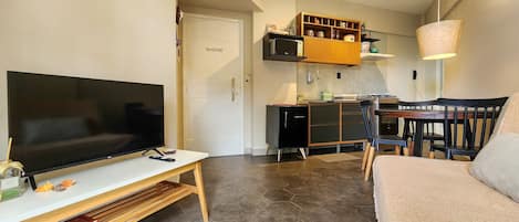 A compact living space with a flat-screen TV on a white stand, a wooden coffee table, a sofa, a dining area with a table and chairs, and a kitchen area with cabinets and a microwave.