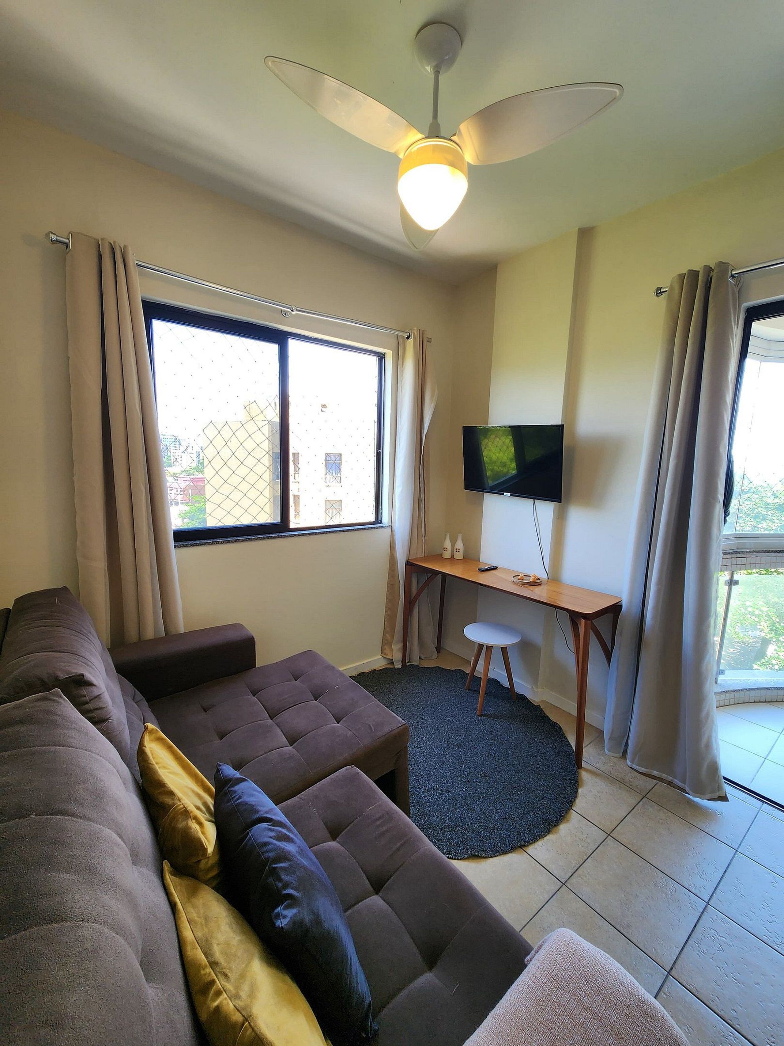 Un salon avec un canapé, un bureau avec une télévision, un ventilateur de plafond et de grandes fenêtres.