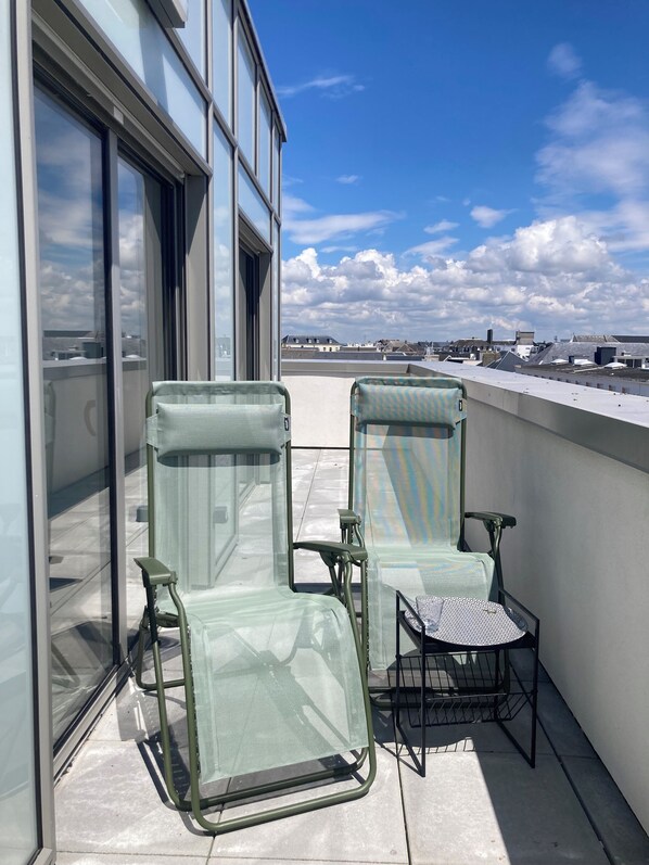 A balcony with two green mesh lounge chairs and a small table, overlooking a cityscape with buildings and a clear sky.