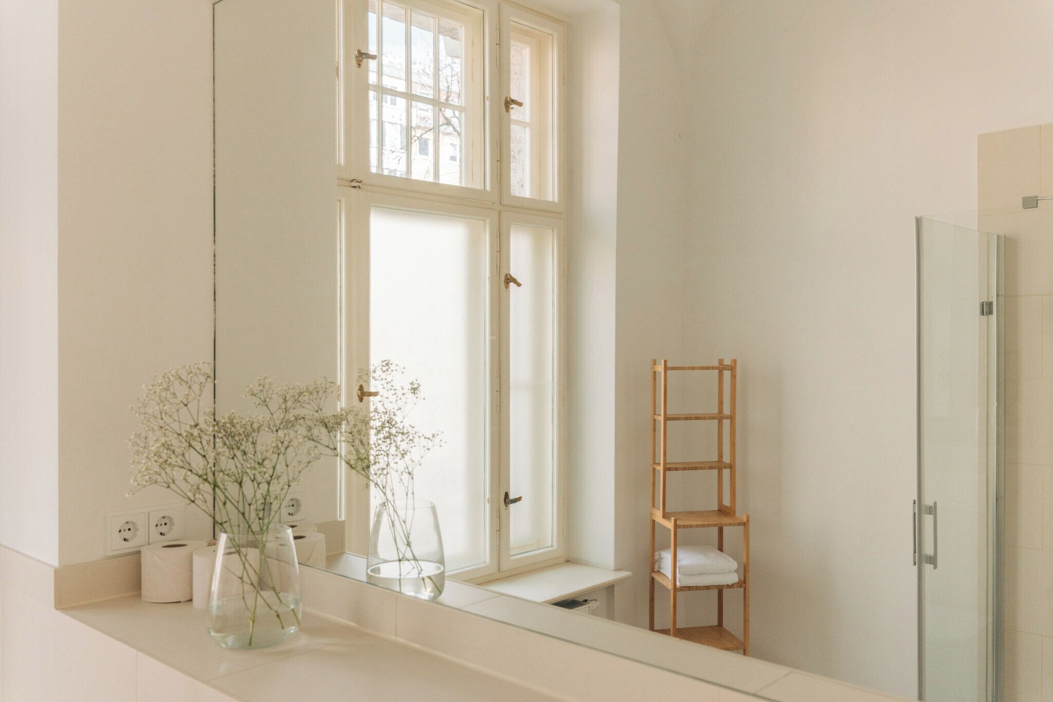 A bathroom with a large mirror, a window, a wooden shelf, and a shower area.