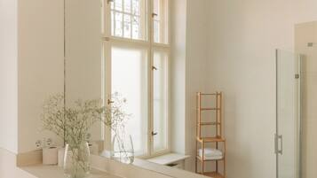 A bathroom with a large mirror, a window, a wooden shelf, and a shower area.