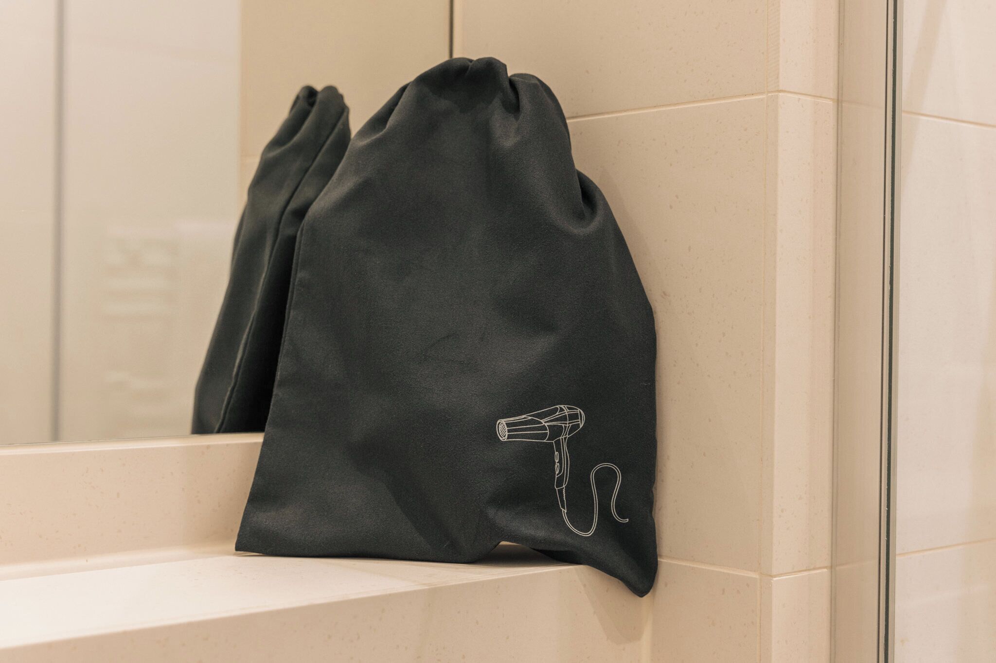 Uma sacola preta com cordão e um desenho de secador de cabelo, pendurada em uma parede com azulejos bege.