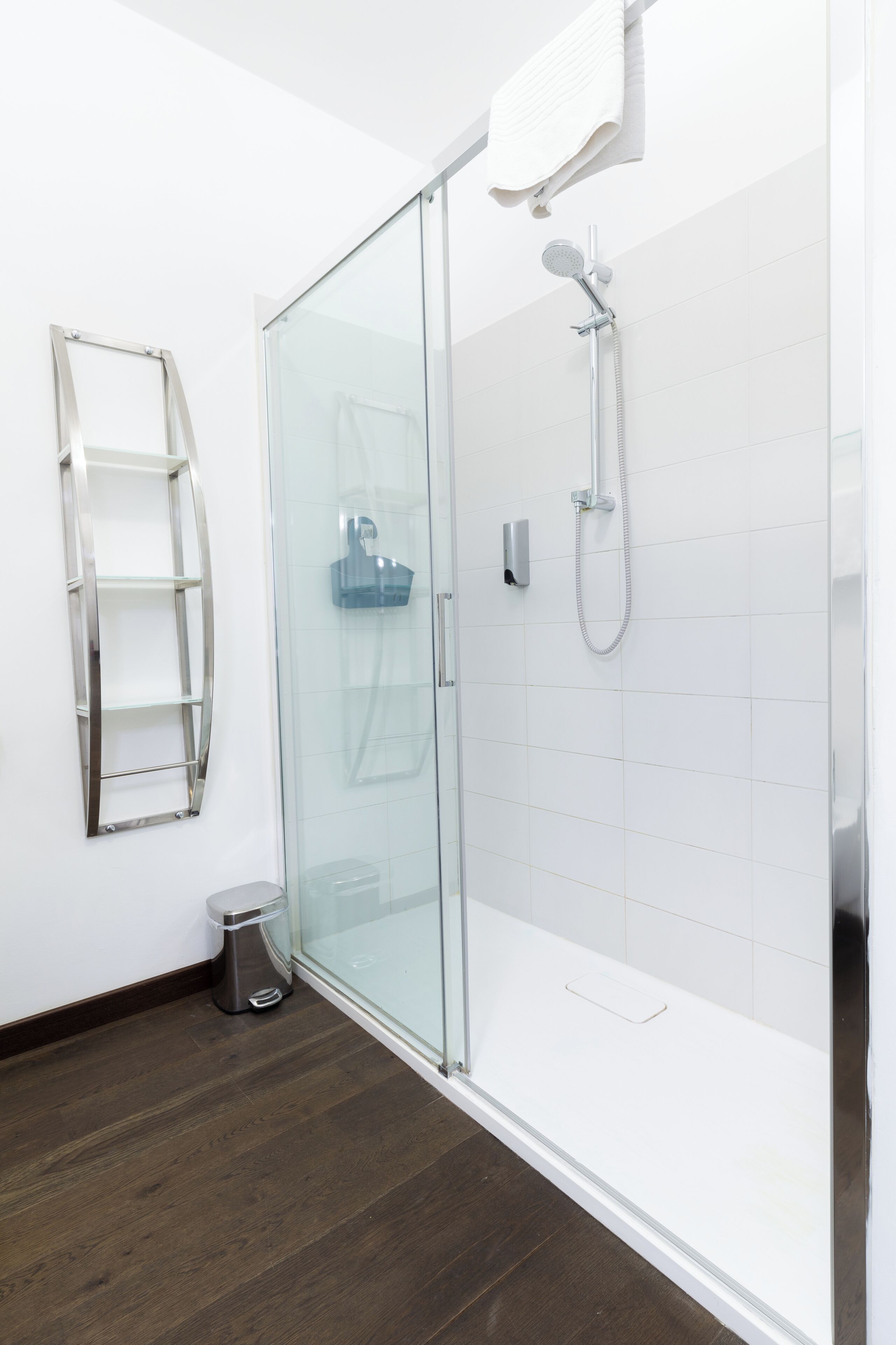 A modern bathroom with a glass shower enclosure, a white tiled wall, a wooden floor, and a metal shelf.