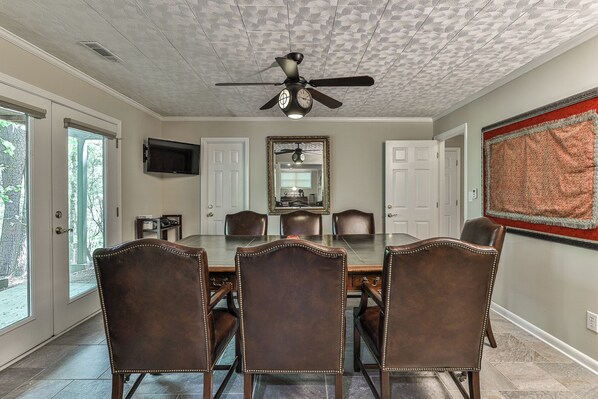 A dining room with a large table, six chairs, a ceiling fan, and a television mounted on the wall.