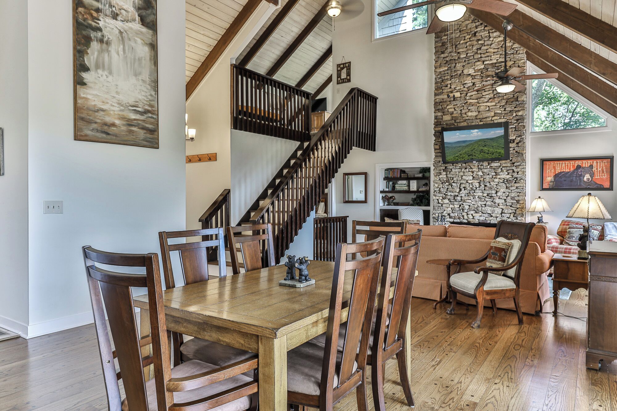 A spacious living area with a wooden dining table, multiple chairs, a staircase with wooden railings, a stone wall, and a large window.