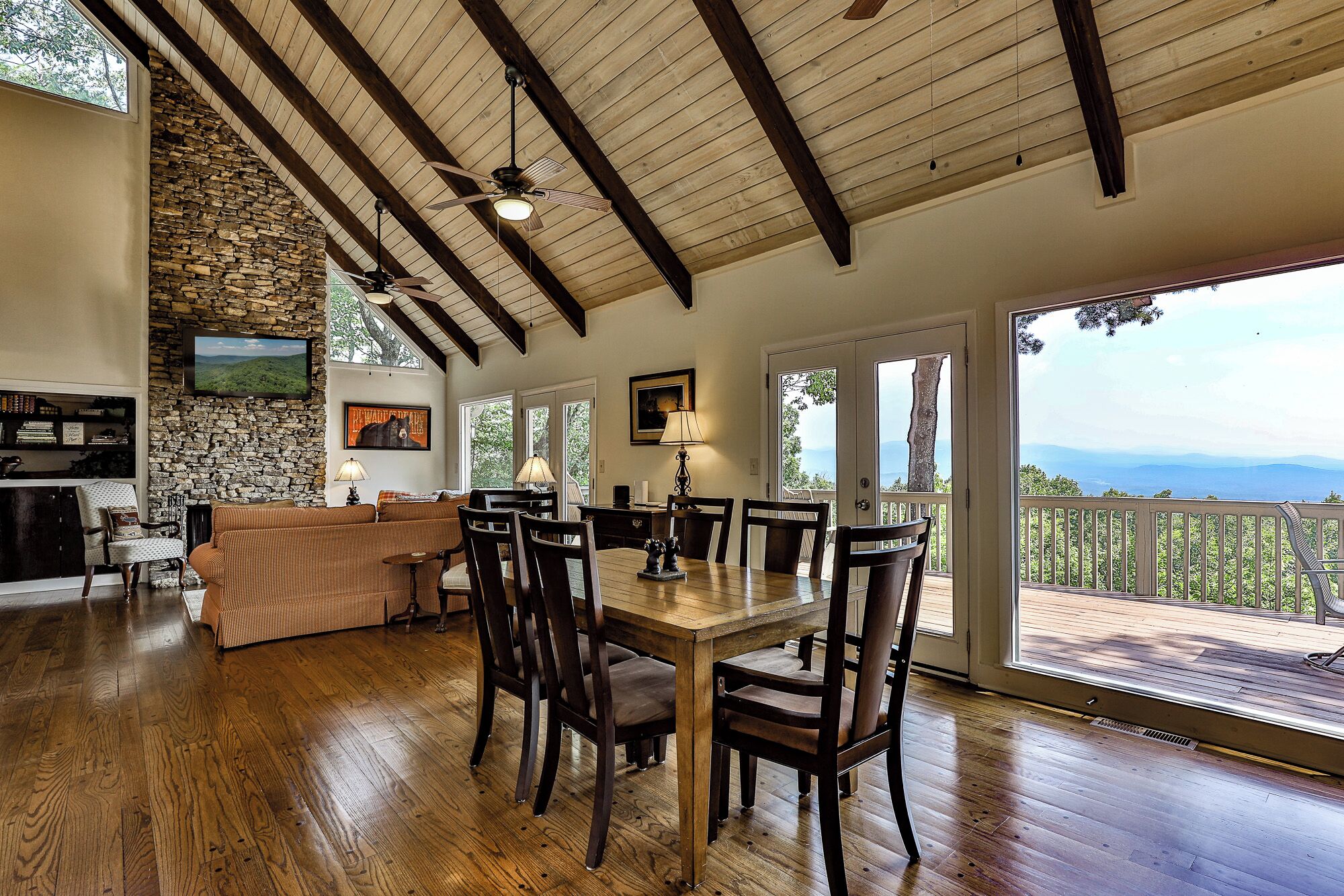 Uma sala de estar ampla com parede de pedra, vigas de madeira no teto, lareira e área de jantar com mesa e cadeiras.