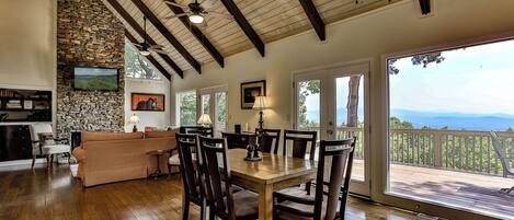 Uma sala de estar ampla com parede de pedra, vigas de madeira no teto, lareira e área de jantar com mesa e cadeiras.