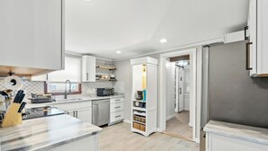 A modern kitchen with white cabinetry, a built-in oven, and a microwave. There is a wooden countertop with a knife block and a dish rack. The floor is light-colored wood, and the kitchen has a bright, airy feel with recessed ceiling lights. - Pool, Firepit, Walk To Beach- A Haven By The Sea (North Myrtle Beach)