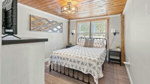 A bedroom with a wooden ceiling, a bed with a patterned bedspread, a wall-mounted TV, and a window with blinds. - Pool, Firepit, Walk To Beach- A Haven By The Sea (North Myrtle Beach)