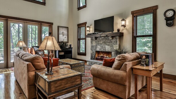 A spacious living room with leather sofas, a stone fireplace, and large windows offering a view of the outdoors. - Elk Horn - Big Canoe GA (Big Canoe)