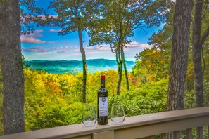 Una botella de vino y dos copas sobre un repisa de madera con una vista panorámica de montañas y árboles.