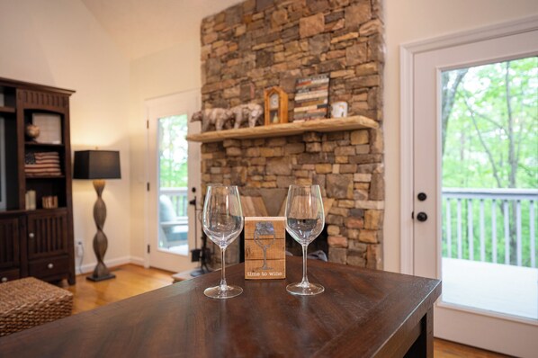 A wooden table with two wine glasses and a card labeled "time to wine" in a room with a stone fireplace, a wooden shelf with decorative items, and a view through a glass door.