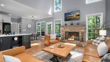 A spacious living room with a fireplace, leather sofas, a coffee table, and large windows offering views of the outdoors.