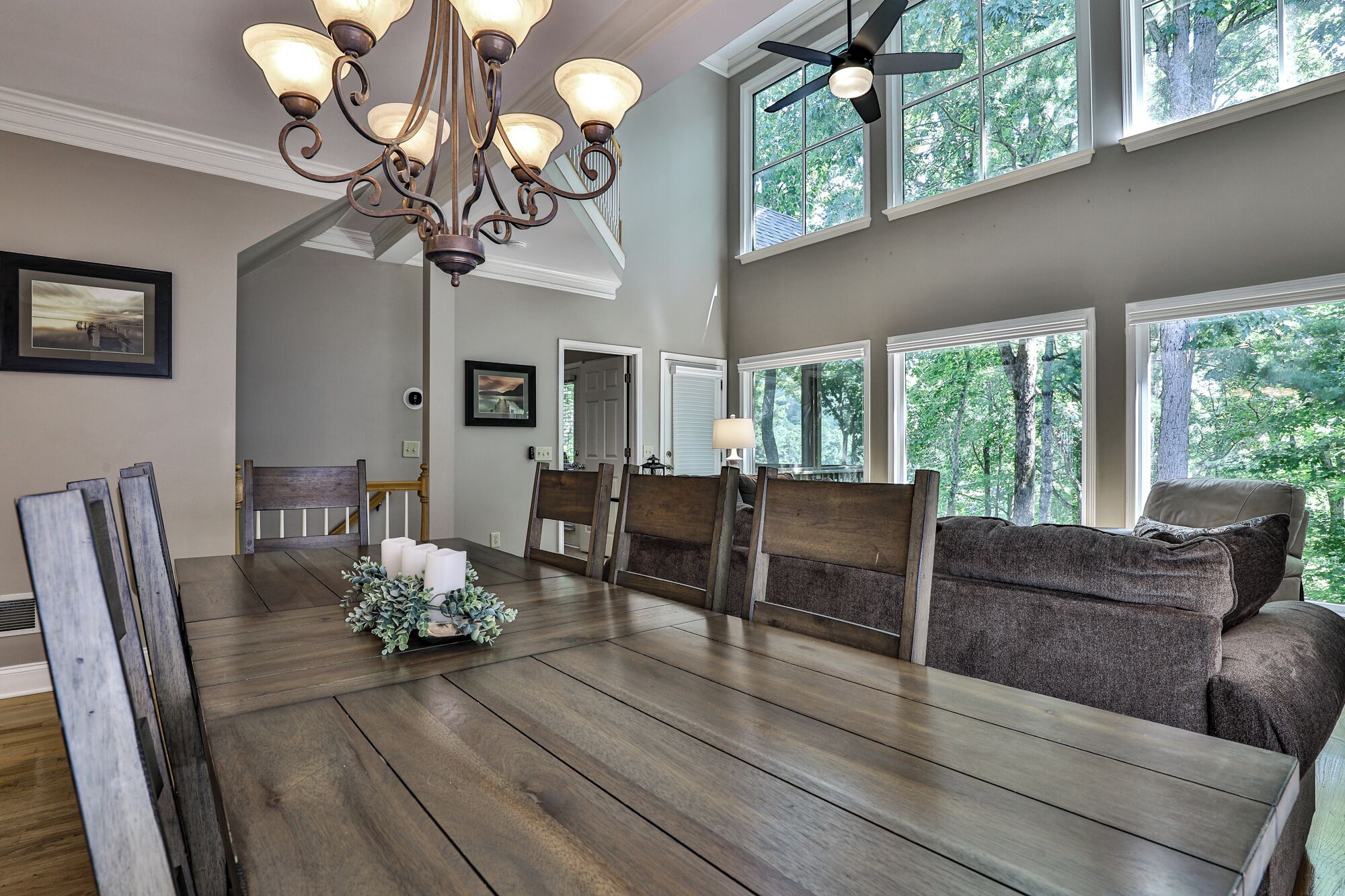 A dining area with a wooden table, chairs, and a chandelier. There are large windows with a view of trees.