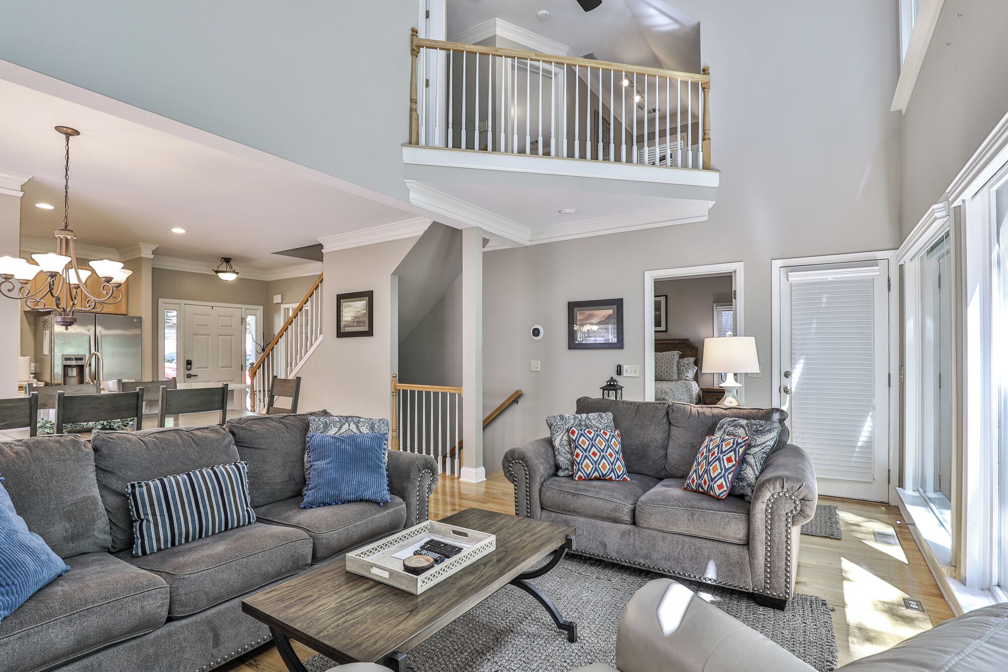 A spacious living room with two grey sofas, a wooden coffee table, and a chandelier. A staircase with a wooden railing leads to an upper level.