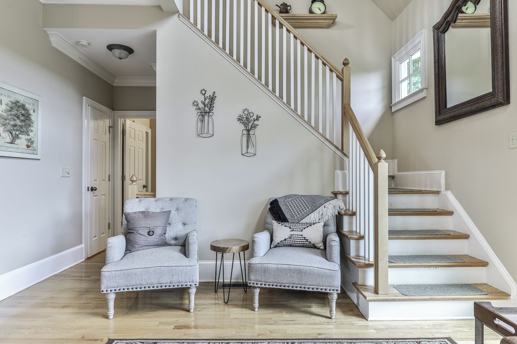 A staircase with a wooden handrail and white balusters, flanked by two grey upholstered armchairs with decorative pillows, a small round table, and a wall-mounted light fixture.