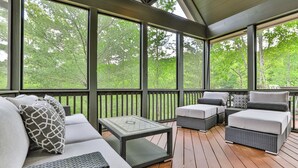 Un área de estar al aire libre techada, con muebles de mimbre y una mesa con cubierta de vidrio, con vista a un área boscosa.