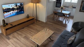 Un salon moderne doté d’un téléviseur à écran plat, d’un meuble de rangement en bois, d’un lampadaire, d’une table basse, d’un canapé et d’un coin repas avec une cuisine en arrière-plan.