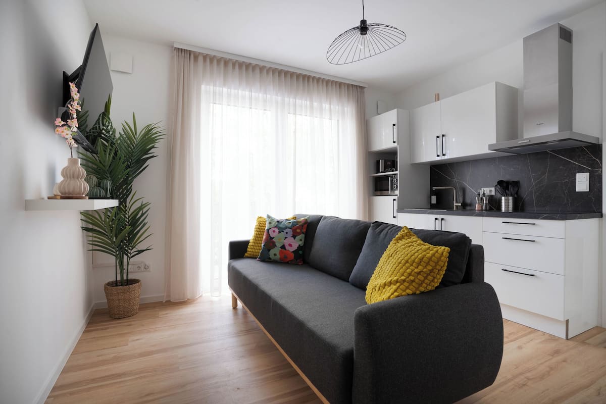 Un salón moderno con un sofá gris, una planta en maceta y una zona de cocina con armarios blancos y encimera negra.