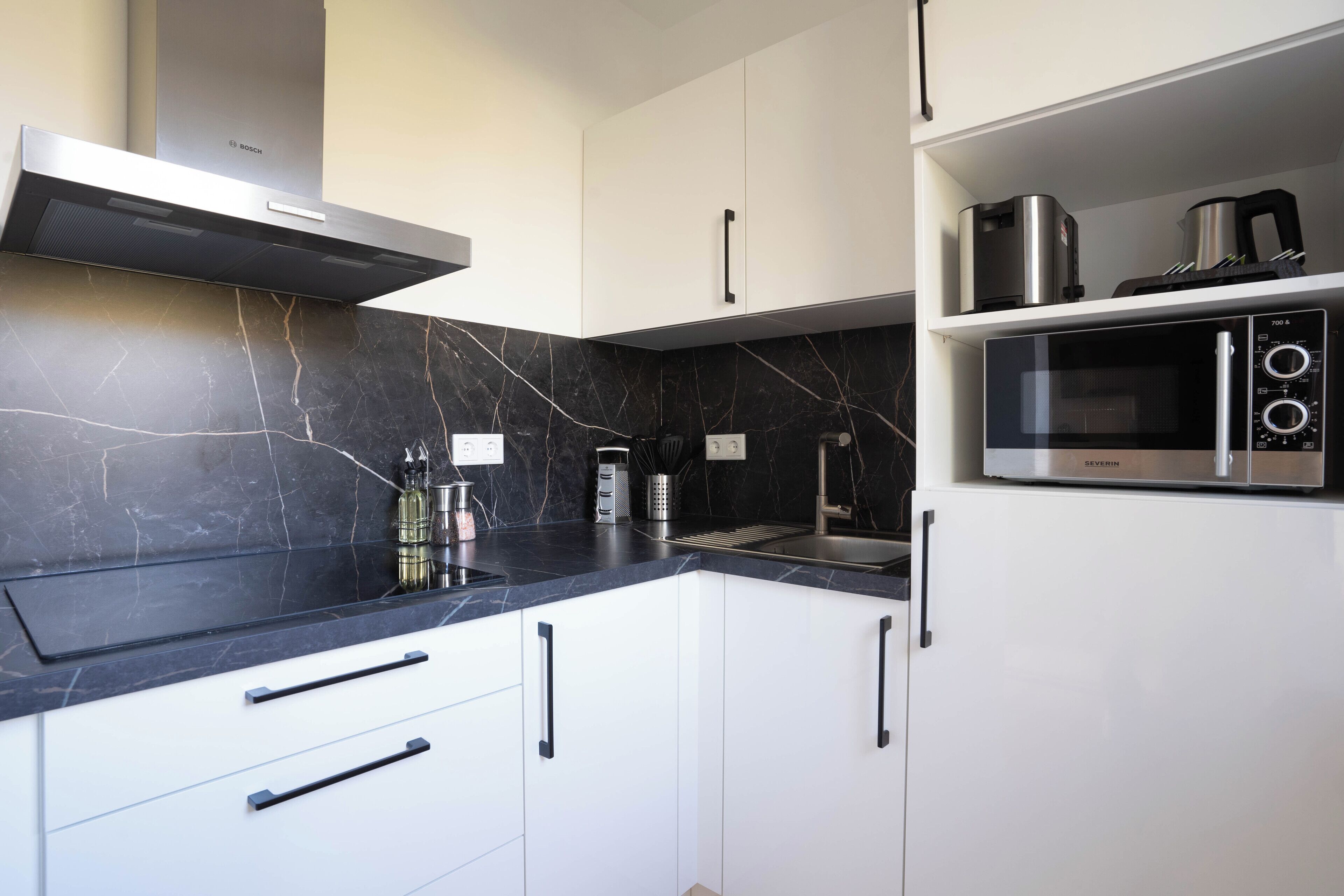 A modern kitchen with white cabinets, a black marble countertop, a Bosch range hood, a Severn microwave, and a coffee maker.