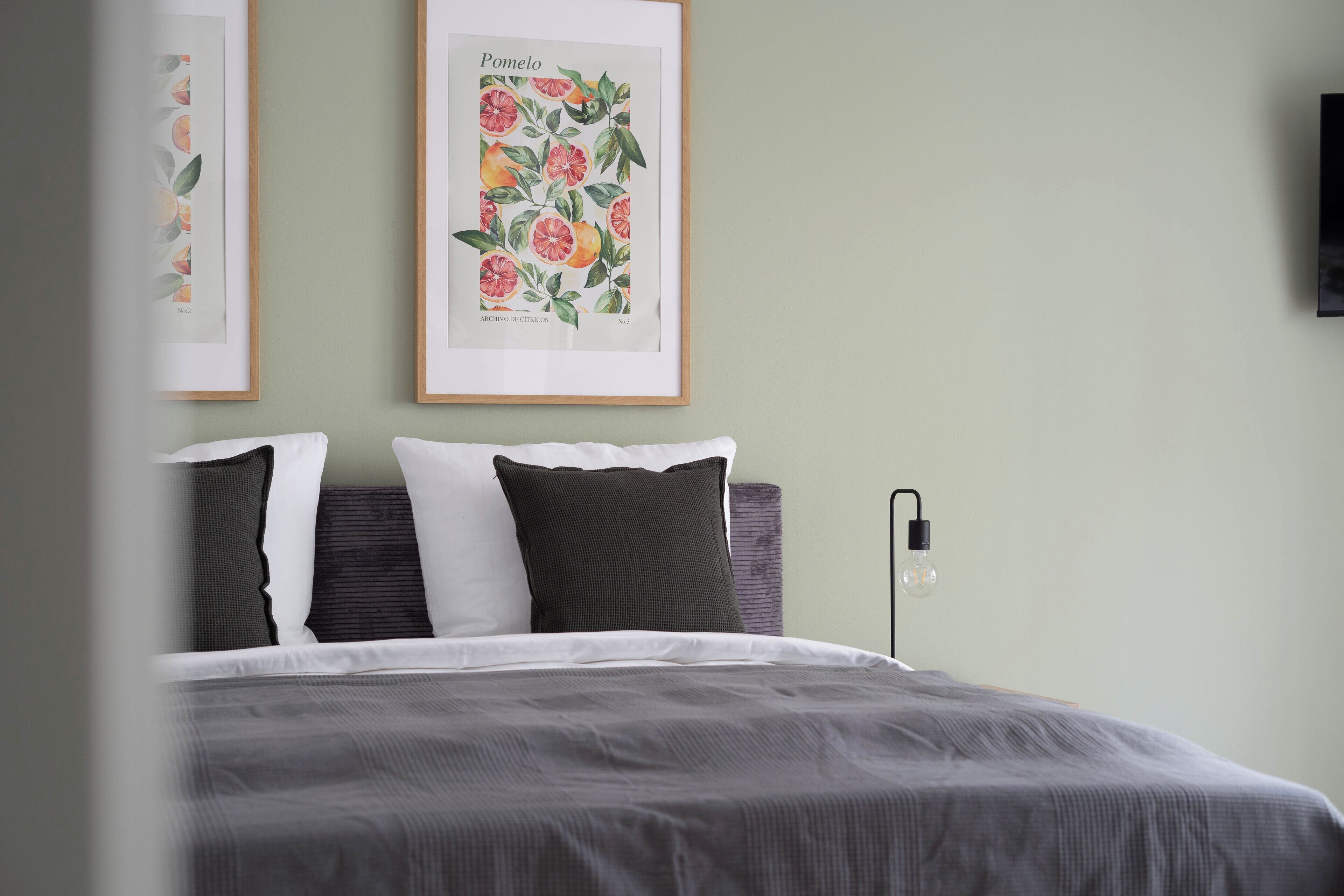 Una cama bien hecha con una colcha gris y almohadas, un grabado botánico enmarcado en la pared y una lámpara de noche.