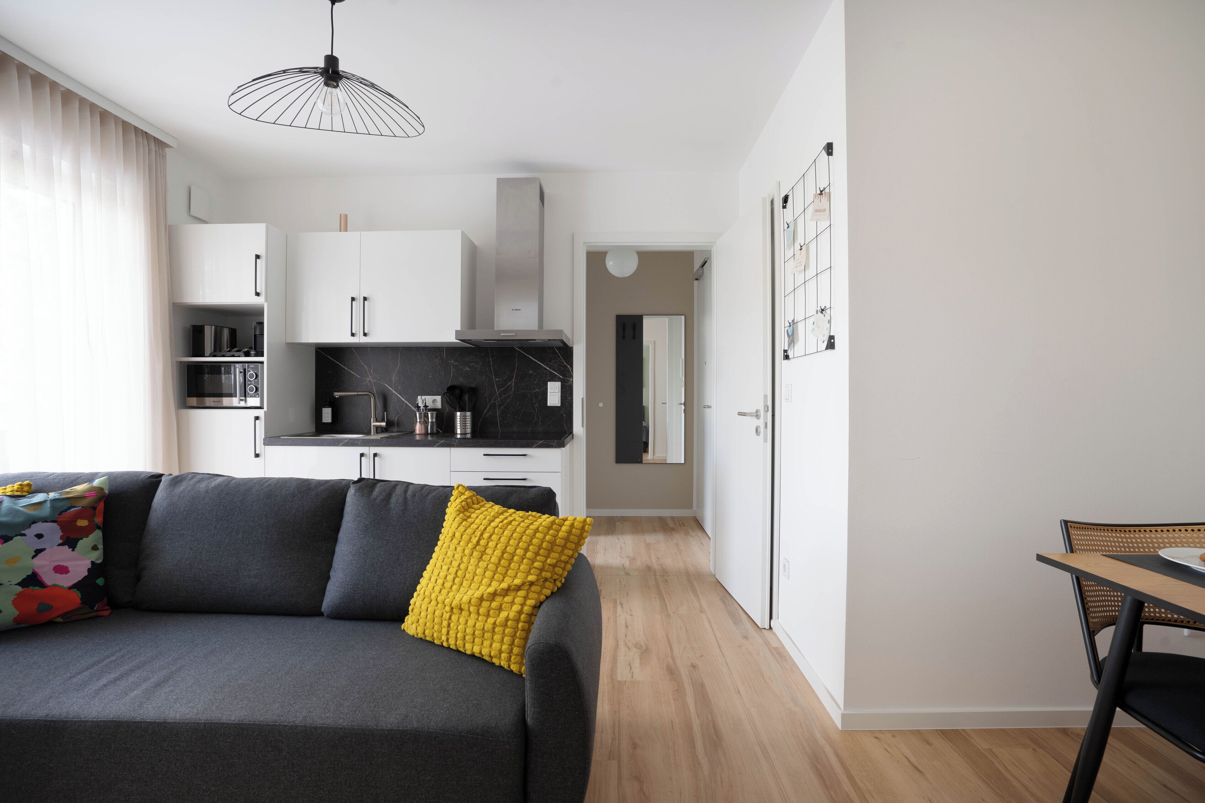 A modern apartment with a grey sofa, a yellow pillow, a dining table, and a kitchen area.