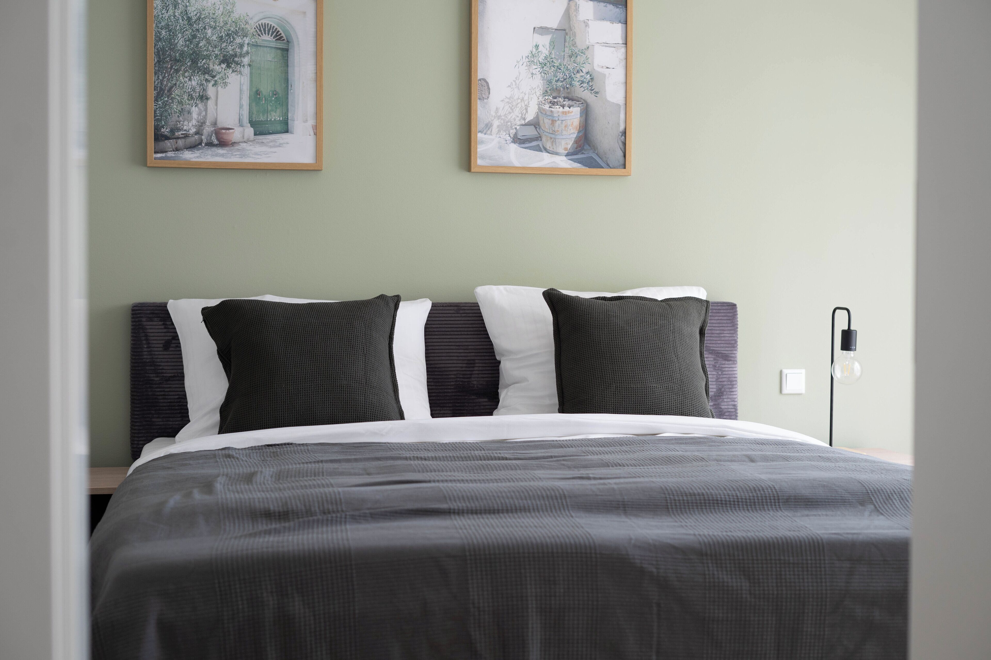 A neatly made bed with a gray and white striped bedspread, two framed pictures on the wall, and a bedside lamp.