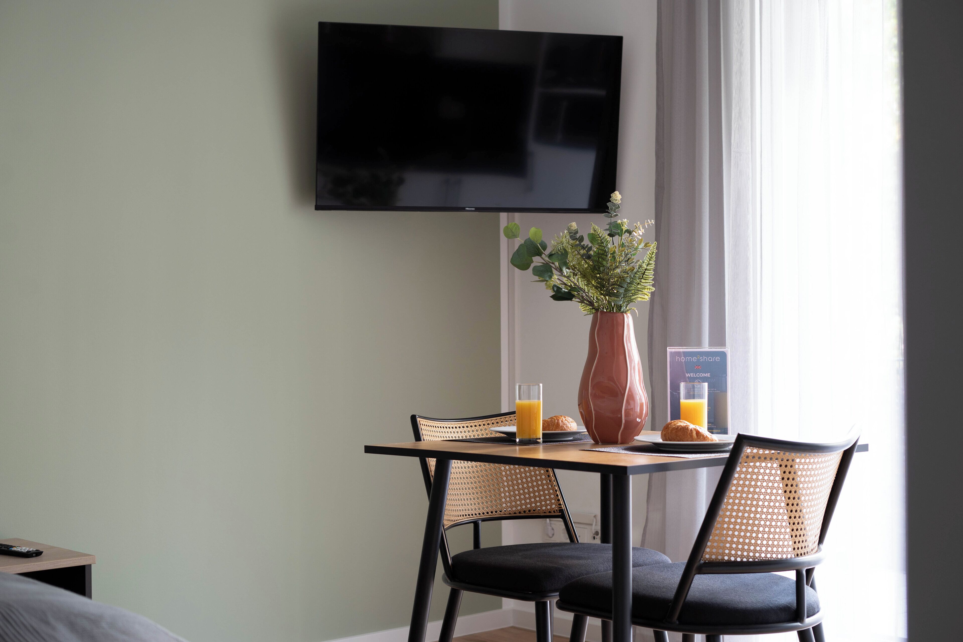 Habitación de hotel con una mesa de comedor, dos sillas, una planta en maceta, un vaso de jugo de naranja, un croissant y un televisor de pantalla plana montado en la pared.