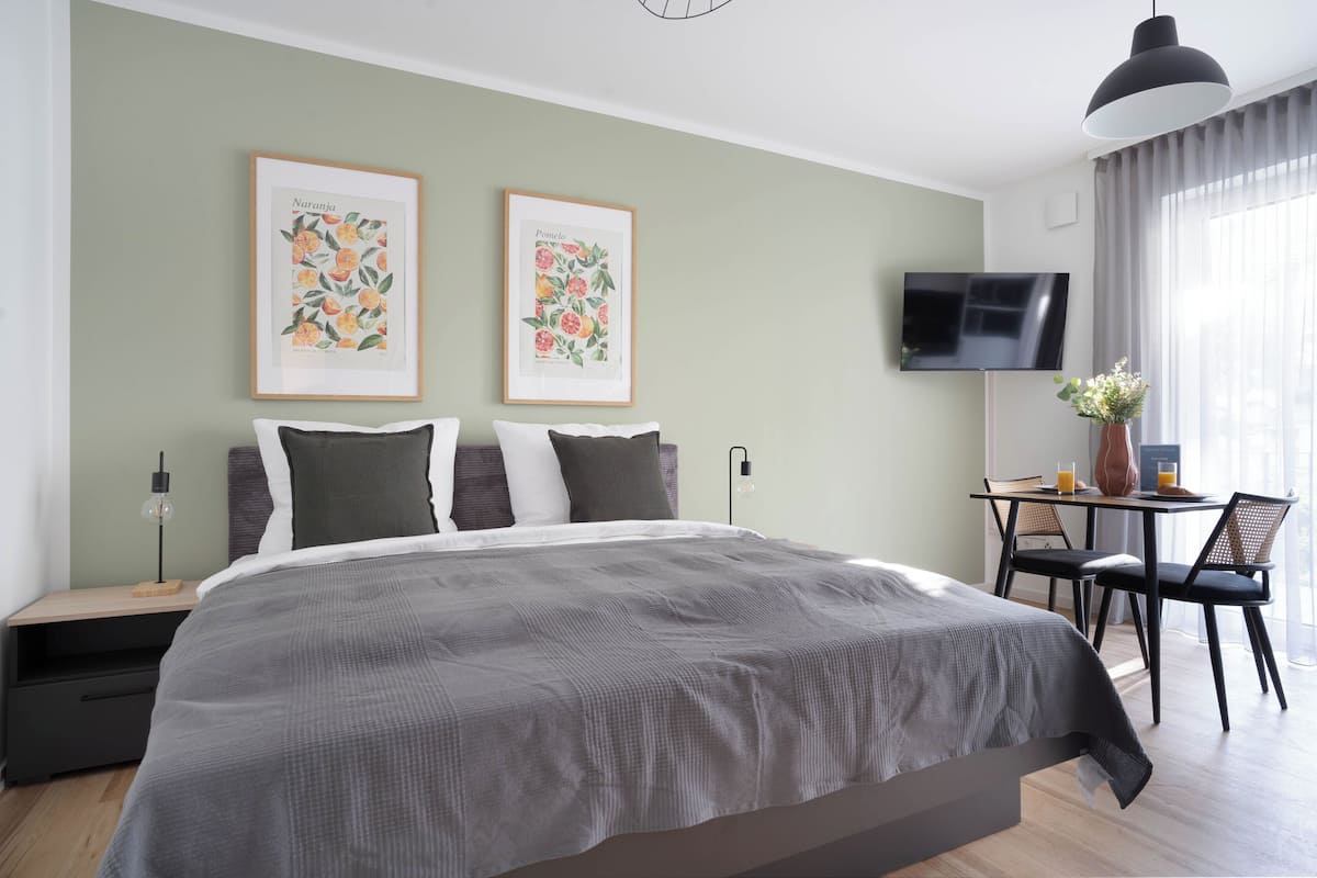 Un dormitorio moderno con una cama grande, un escritorio con sillas, un televisor y dos impresiones enmarcadas de plantas en la pared.