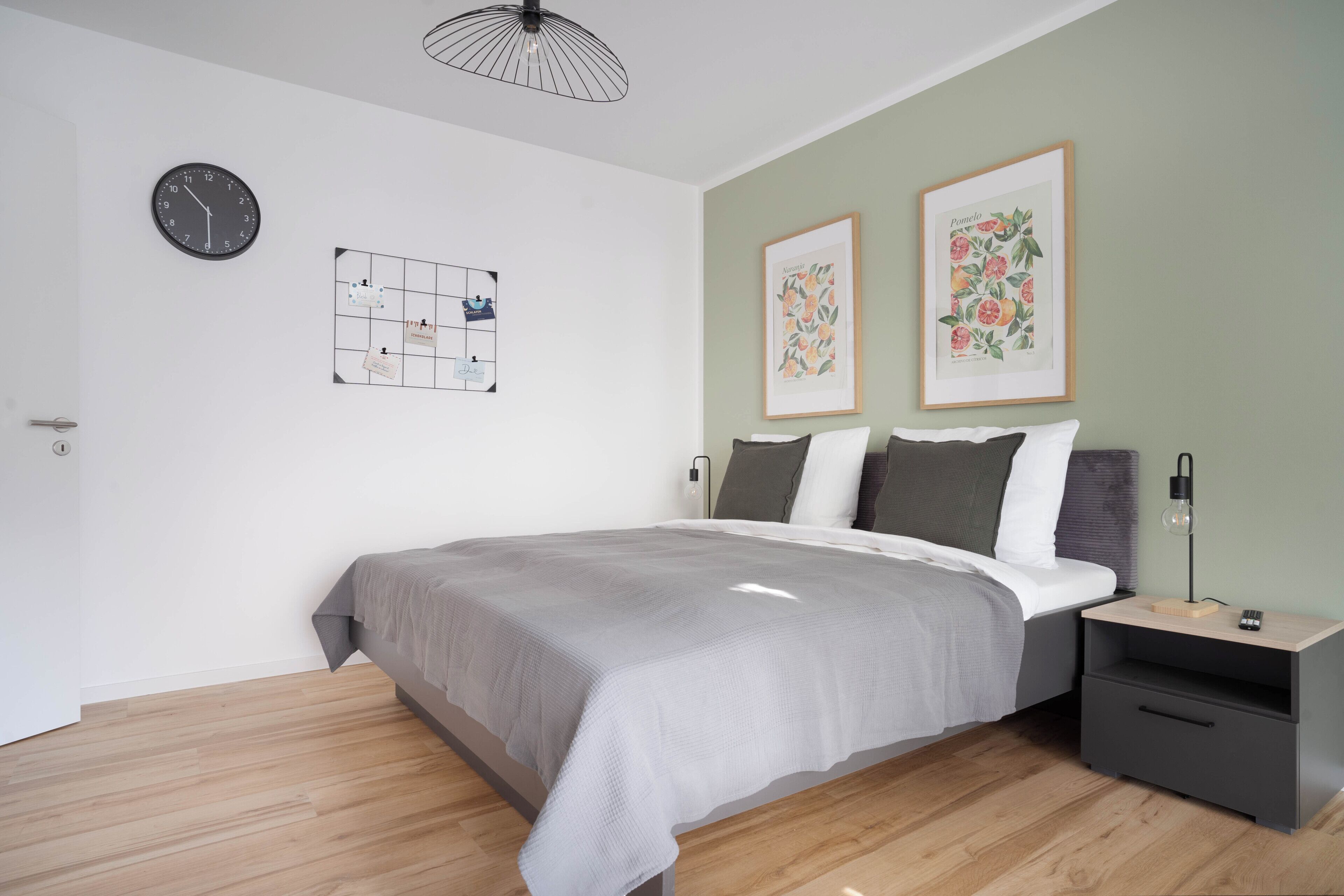Une chambre moderne avec un grand lit, une table de chevet avec une lampe, une horloge murale et deux reproductions botaniques encadrées.