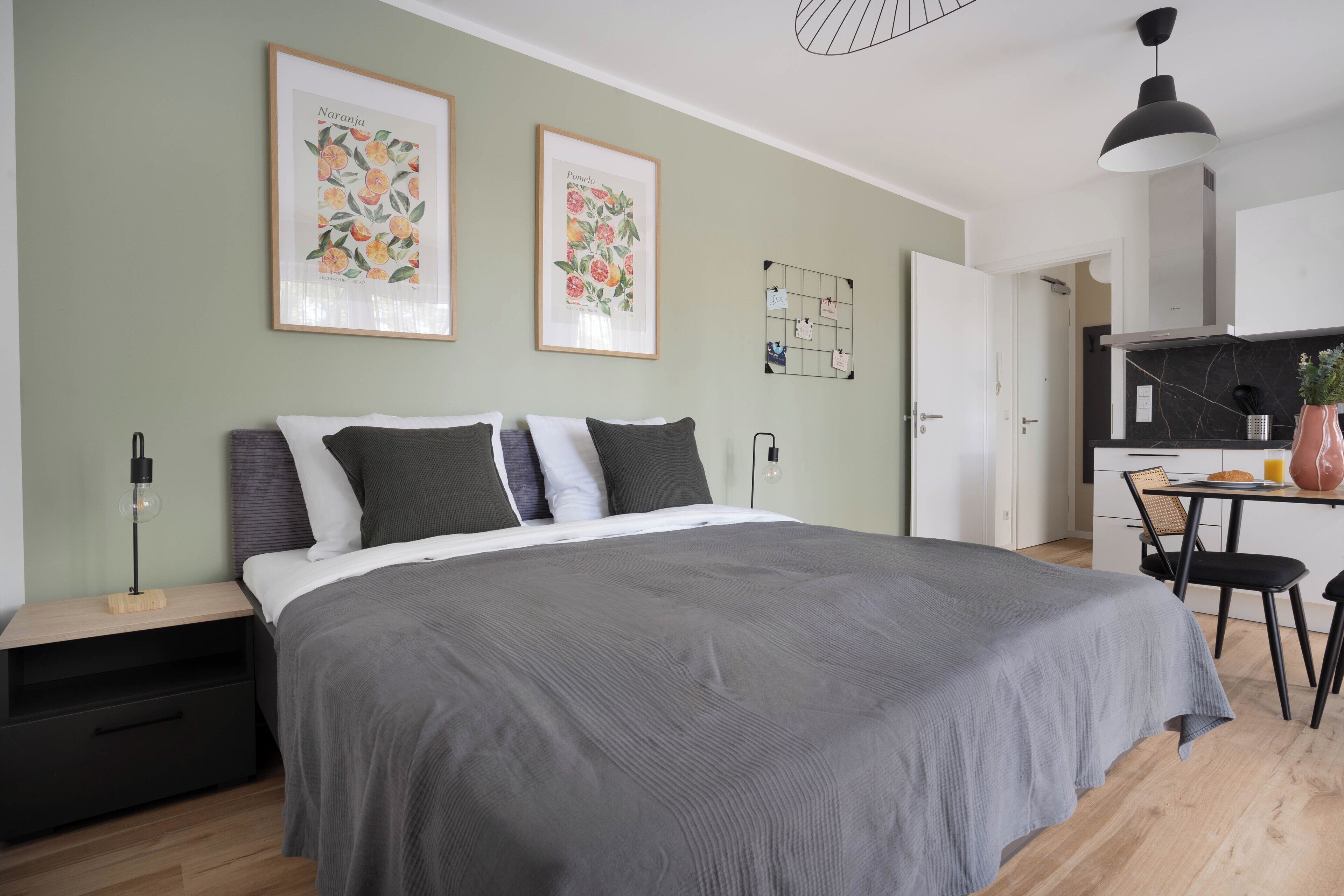 A modern bedroom with a large bed, a bedside table, a nightstand, a desk with a chair, and a kitchen area visible through an open door.