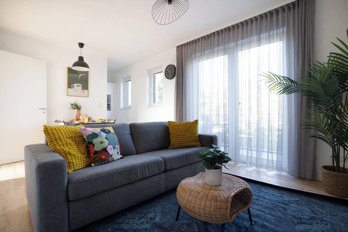 Una sala de estar moderna con un sofá gris, cojines amarillos, una mesa de centro de mimbre y un gran ventanal con cortinas translúcidas.