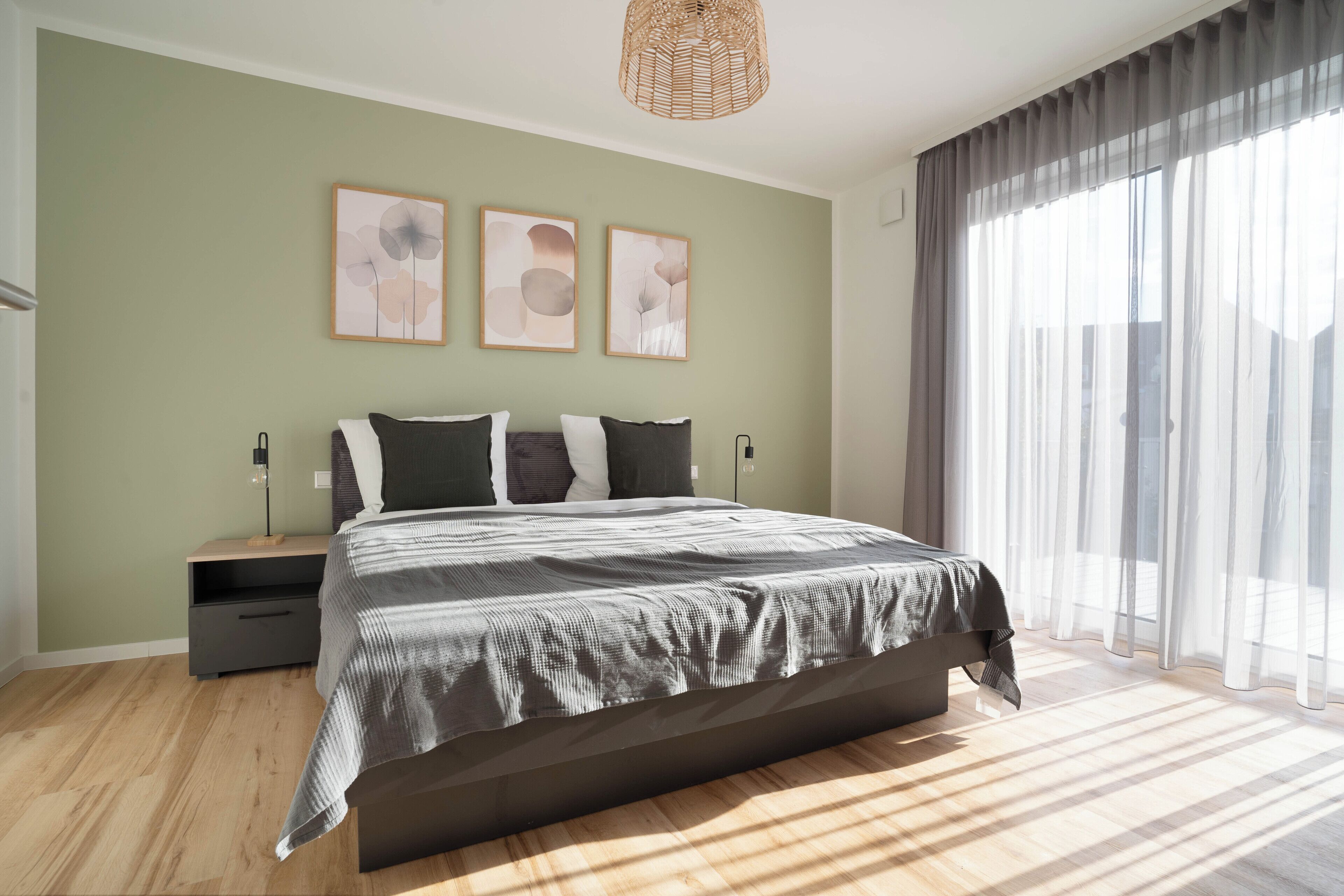 Une chambre à coucher moderne avec un grand lit, une table de chevet et une commode. Trois tableaux abstraits ornent le mur, et une grande fenêtre est agrémentée de rideaux légers.