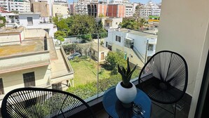 Un balcón con dos sillas negras y una mesa pequeña, con vista a un paisaje urbano de varios edificios y un cielo azul despejado.