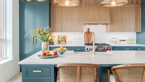 Una cucina con mobili color petrolio, un piano di lavoro bianco, un fornello a gas e due sedie in vimini.