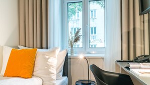 A hotel room with a bed, a desk, a chair, a window with curtains, and a vase with dried plants.