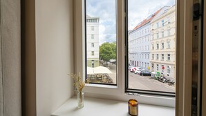 Een raam met uitzicht op een straat, een vaas met gedroogde bloemen en een kaars op de vensterbank.