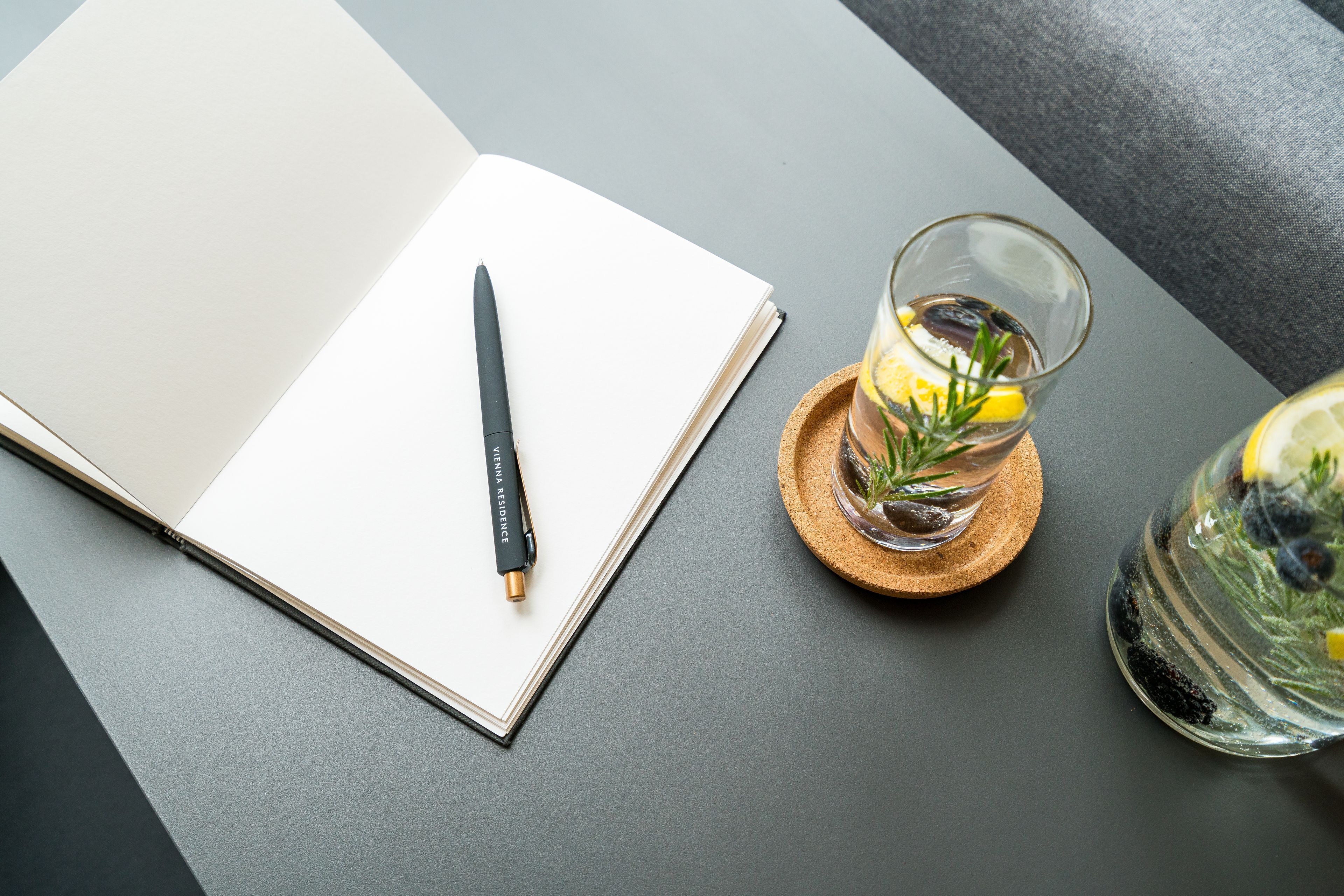 Un cuaderno abierto con un bolígrafo sobre una superficie oscura, un vaso con limón y romero, y un tarro de cristal con un limón.
