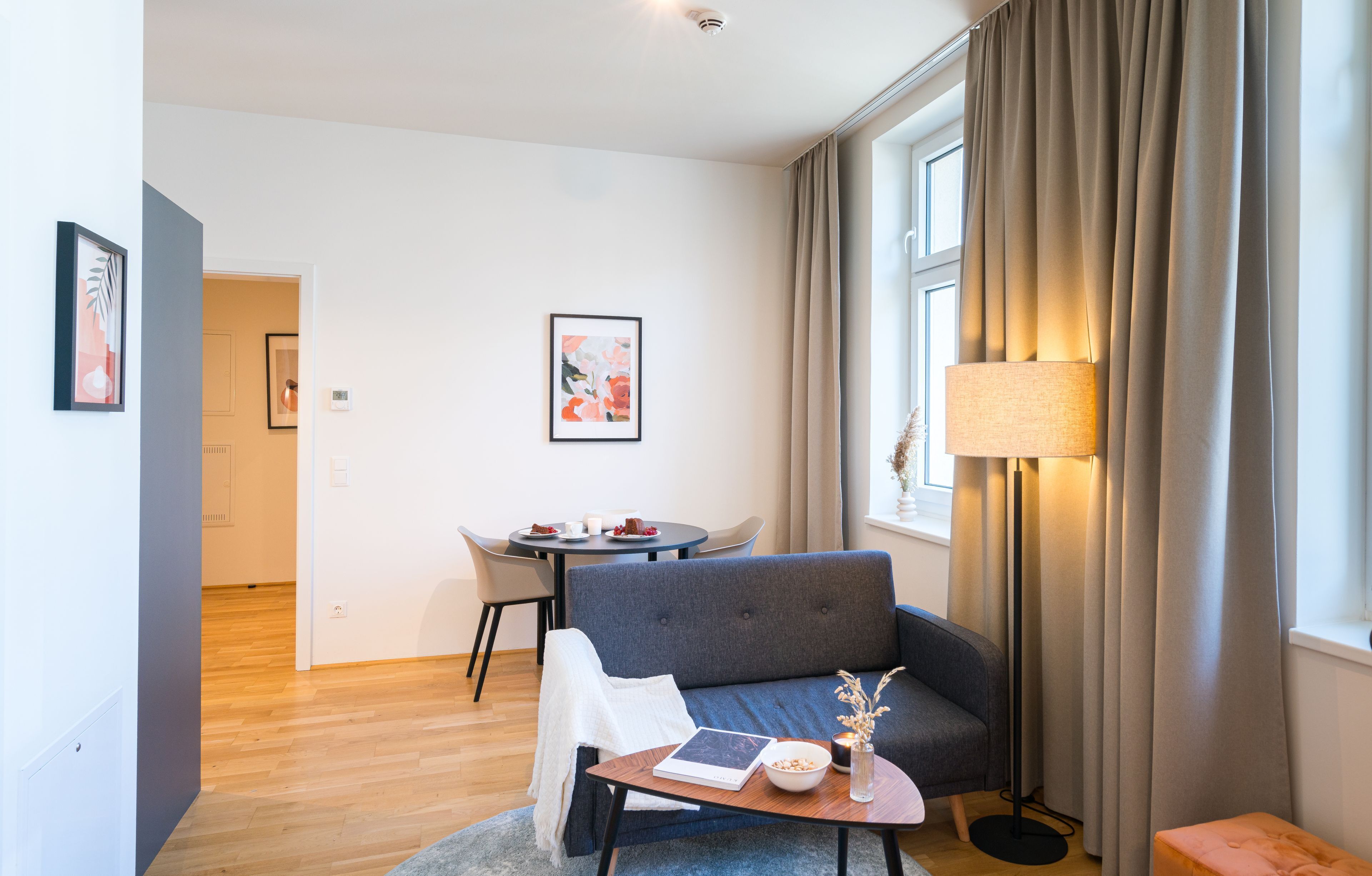 A modern living room with a grey sofa, a round coffee table, a floor lamp, and a dining area with a table and chairs.