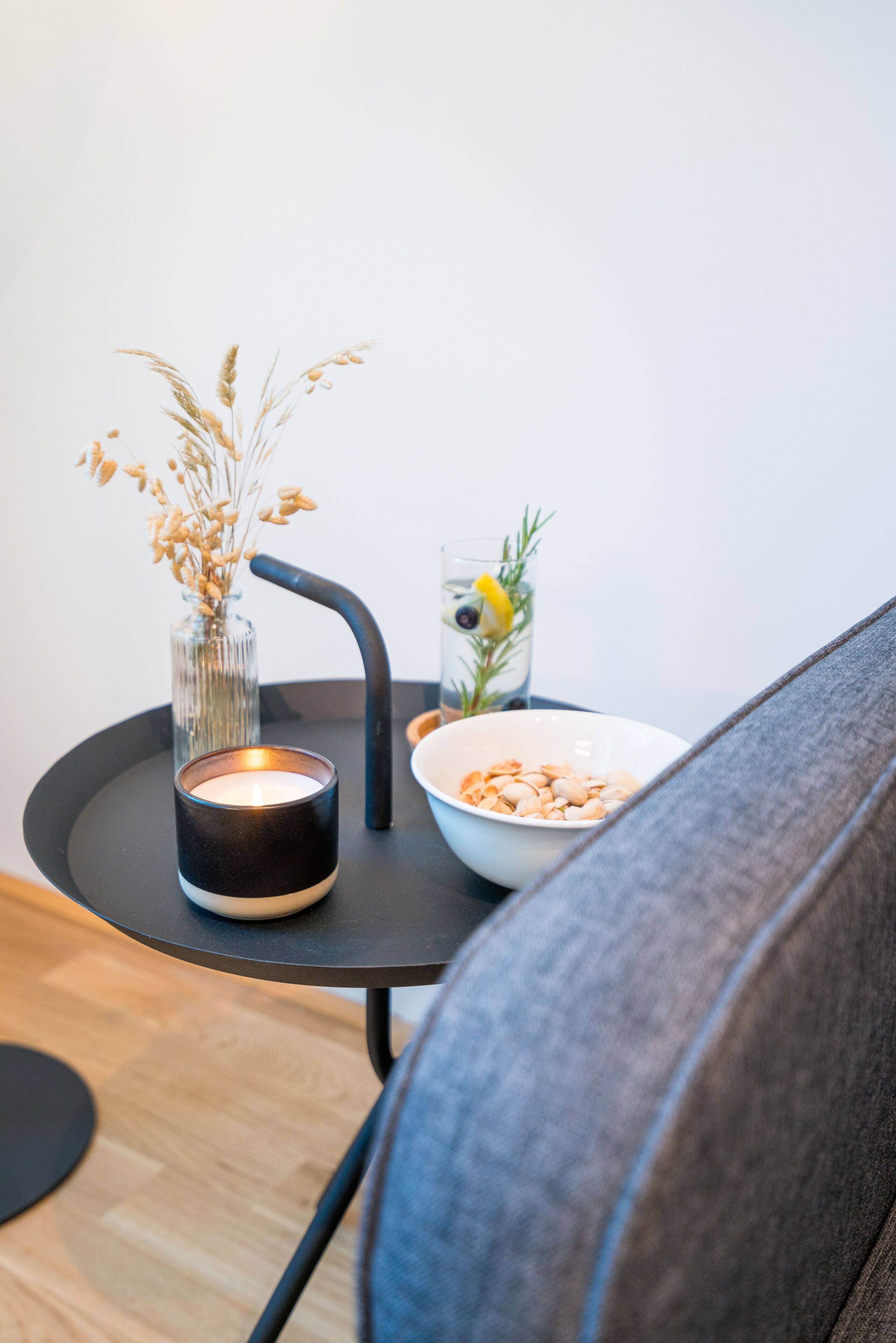 Une petite table ronde avec une bougie, un vase en verre contenant des fleurs séchées, un bol de noix et un verre d’eau avec une tranche de citron et une branche de romarin.