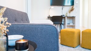 Moderni olohuone, jossa on harmaa sohva, keltainen jalkarahi, valkoinen kulho ja sytytetty kynttilä pöydällä.