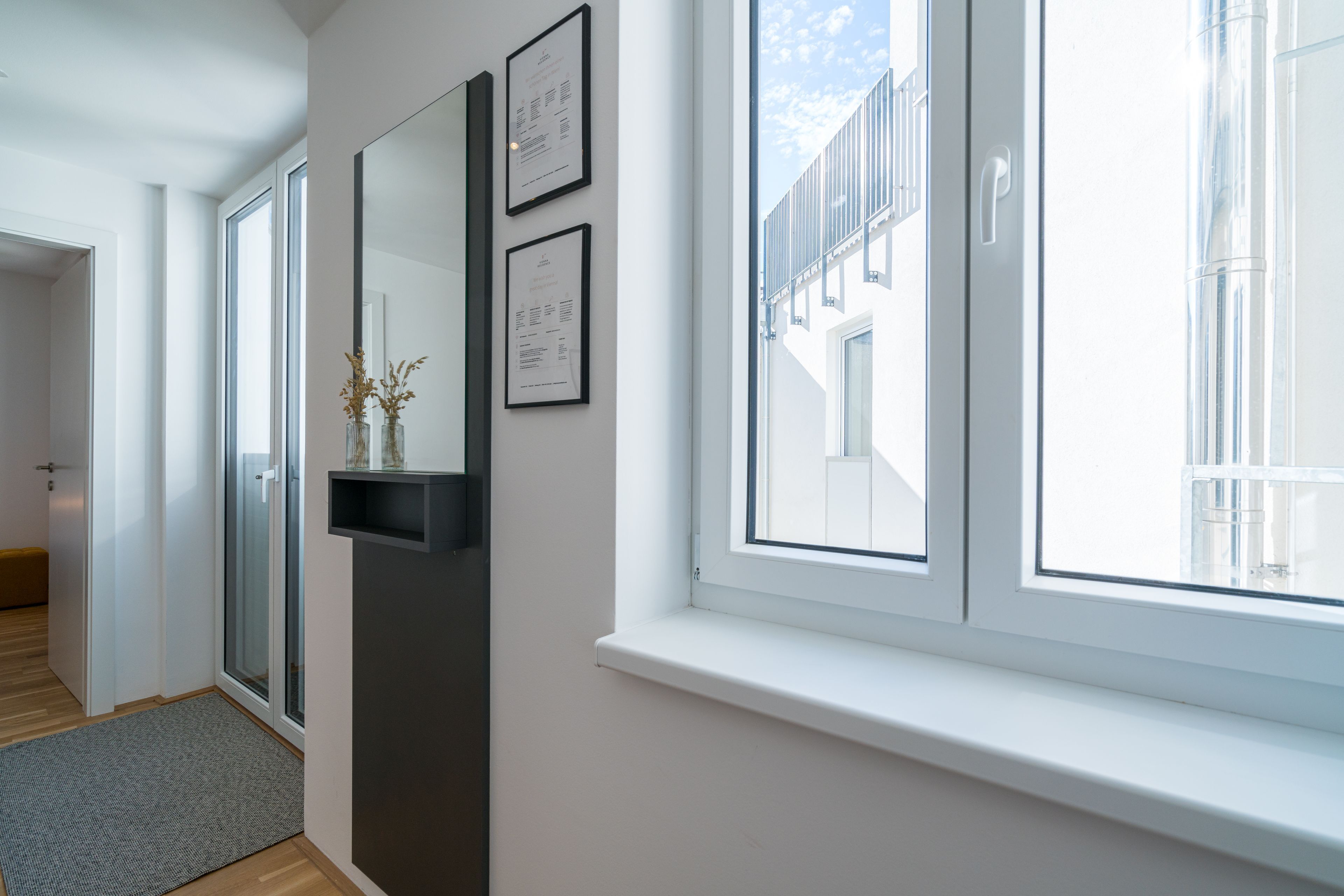 A modern hotel room with a large window, a door leading to a balcony, and a small shelf with a vase.
