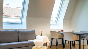 Un salon moderne avec un canapé gris, une petite table entourée de chaises et une fenêtre donnant sur des immeubles.