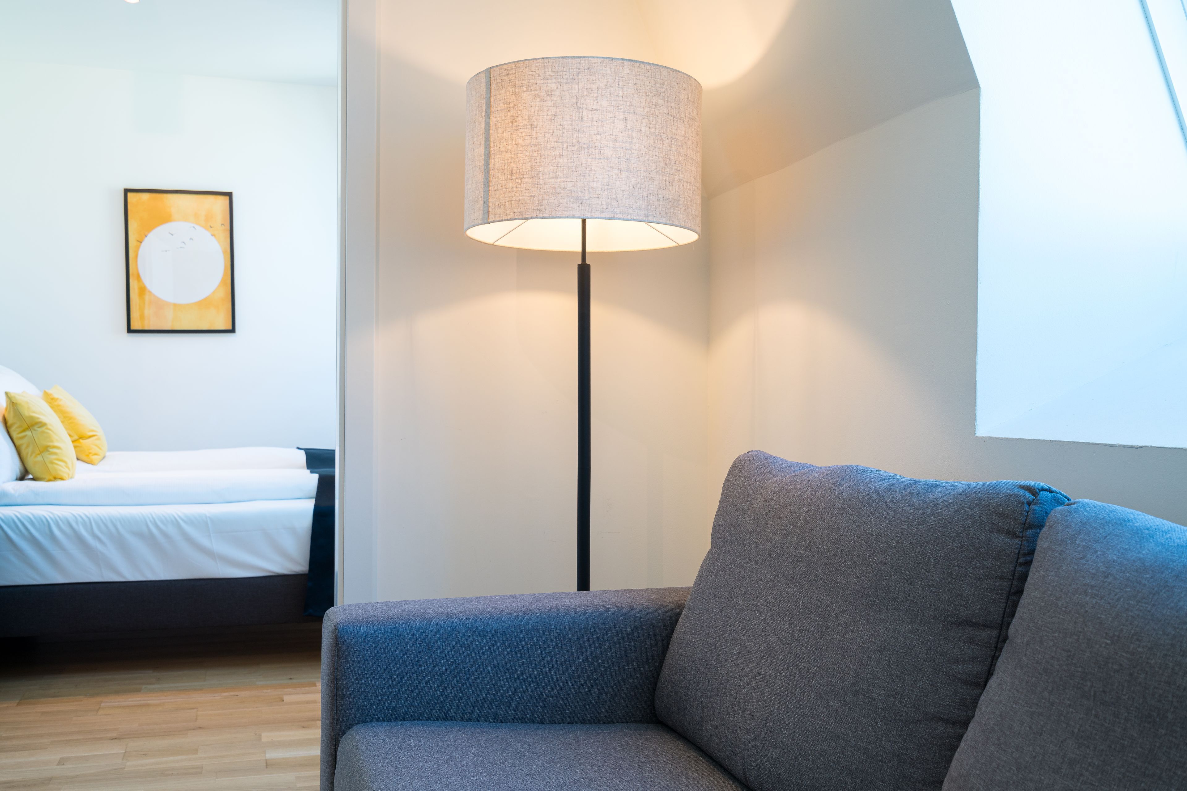 Een moderne kamer met een grijze bank, een vloerlamp, een bed met witte beddengoed en een lijstwerk kunstwerk aan de muur.