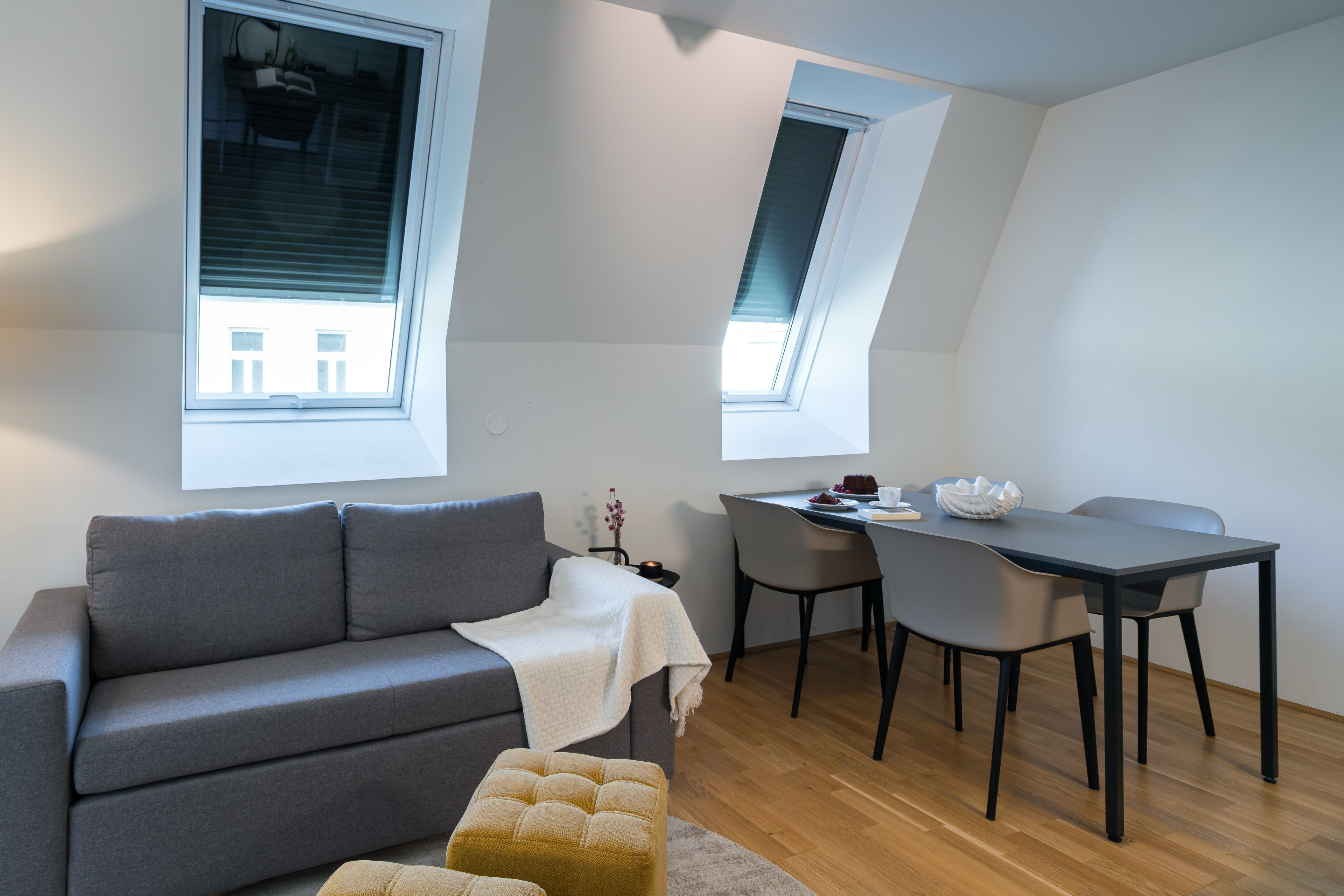 A modern living room with a grey sofa, a small ottoman, a dining table with chairs, and a skylight.
