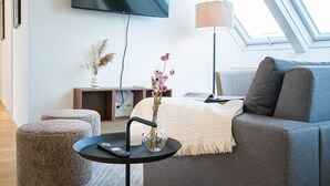 Un salon moderne avec un canapé gris, une petite table ronde sur laquelle se trouve une télécommande, un vase avec des fleurs et un téléviseur à écran plat fixé au mur.